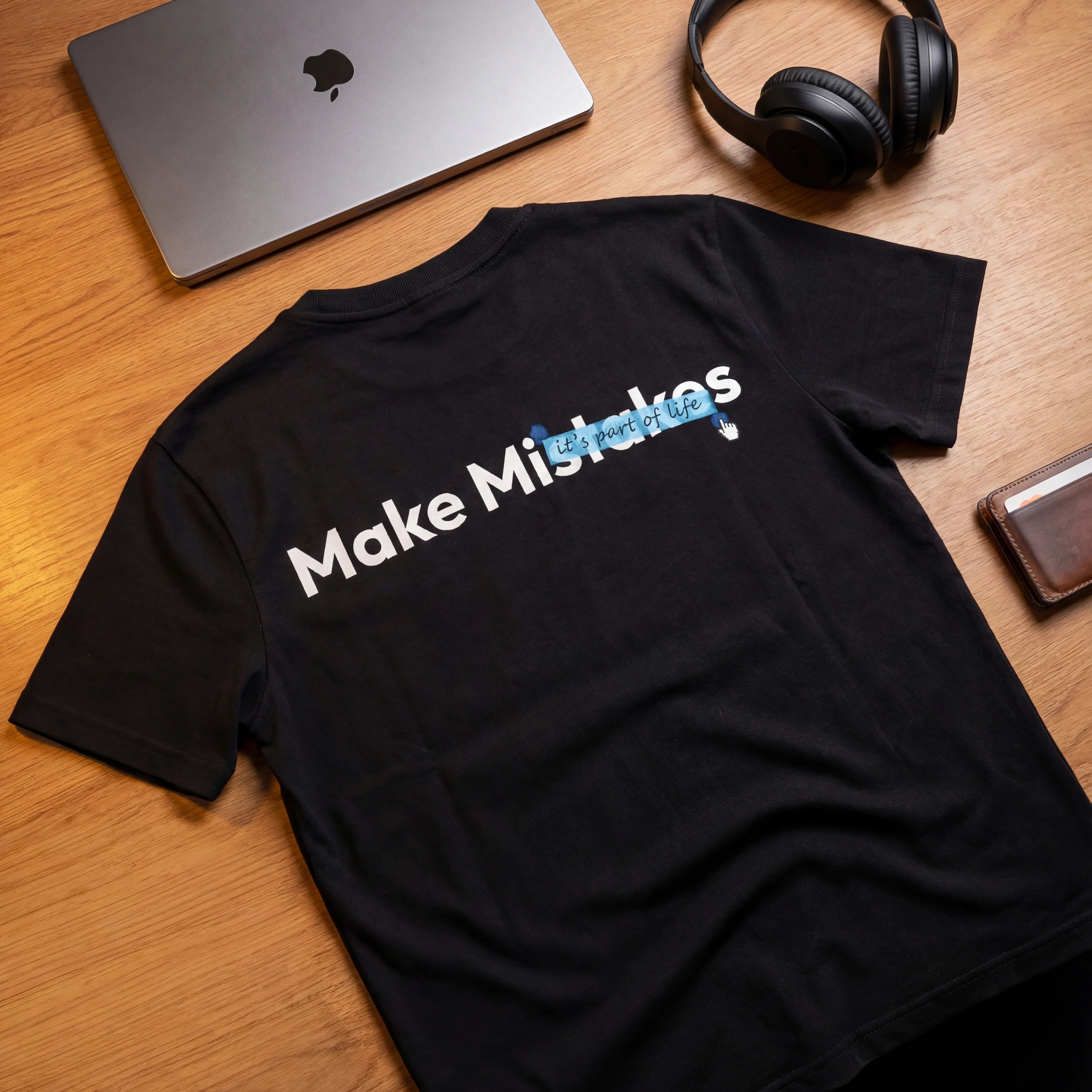Black t-shirt with 'Make Mistakes' text on a wooden surface with laptop, headphones, and wallet.