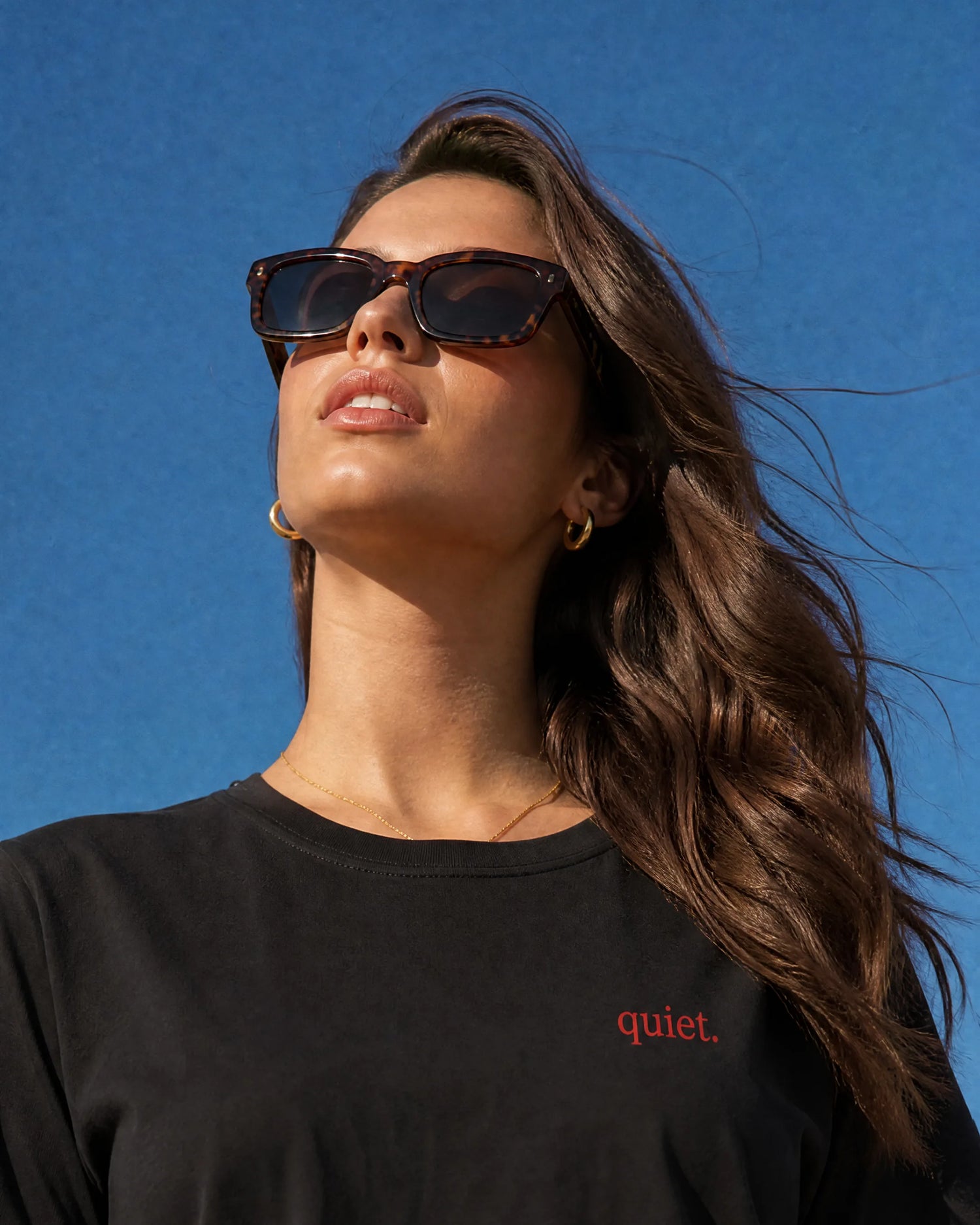 Zehra wearing sunglasses and a black t-shirt from zeromode with 'quiet.' text on the front against a blue sky
