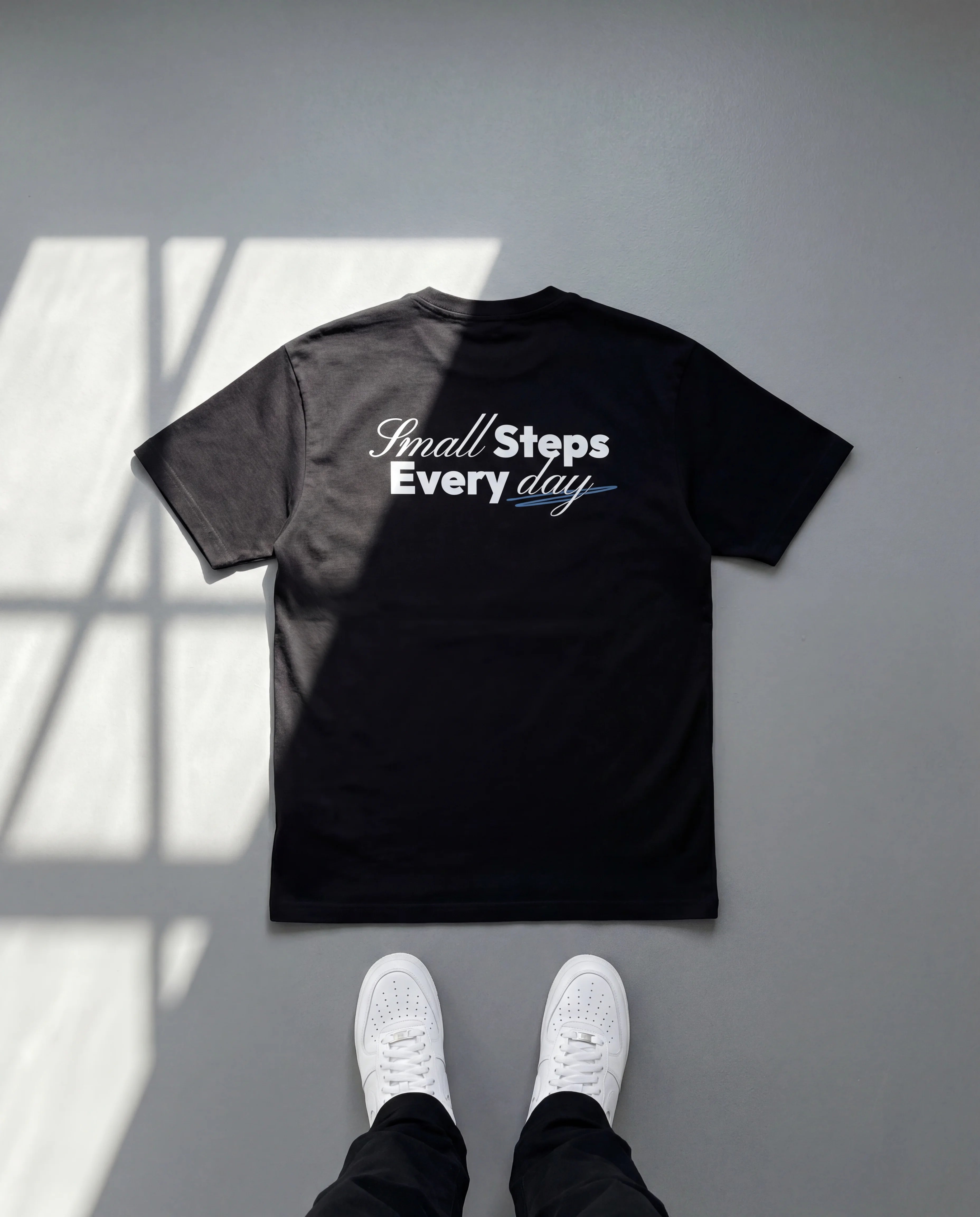 Black t-shirt with 'Small Steps Every Day' text on a gray floor with white shoes.