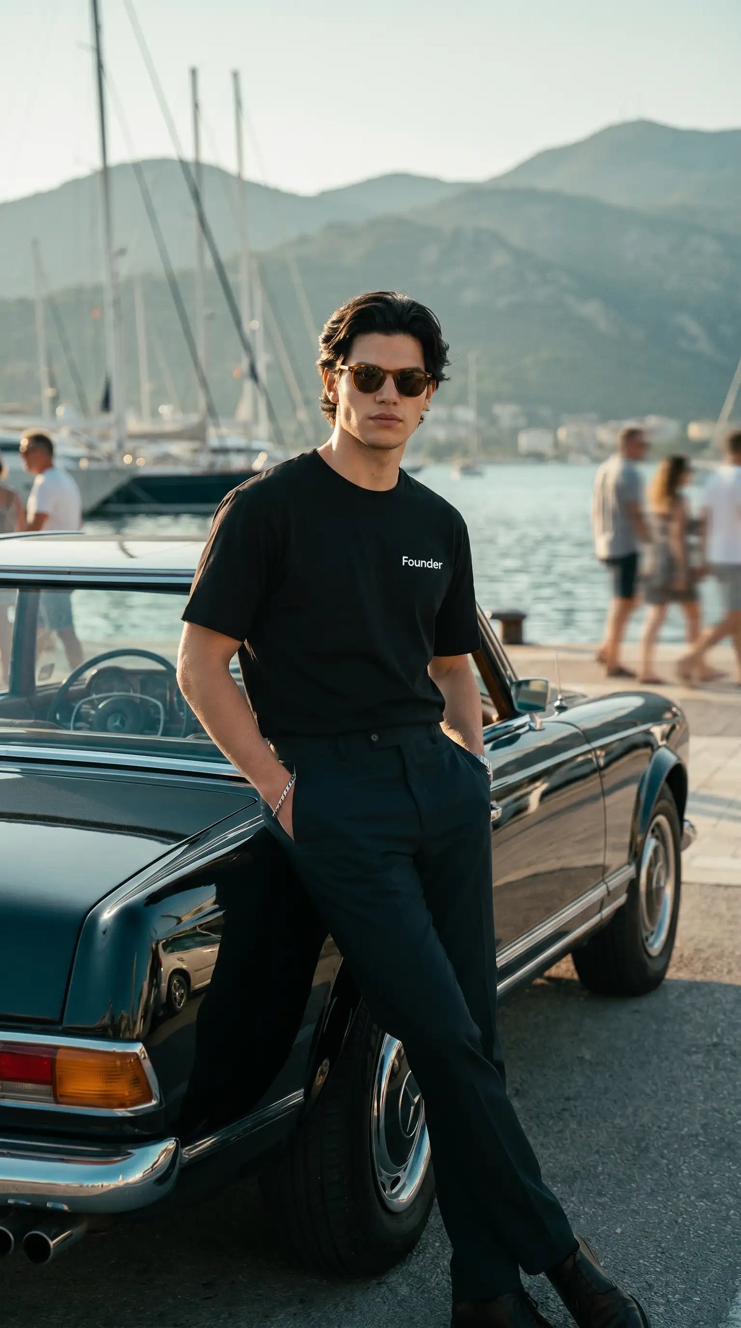 Man in sunglasses and black clothing leaning against a classic car with a marina and mountains in the background