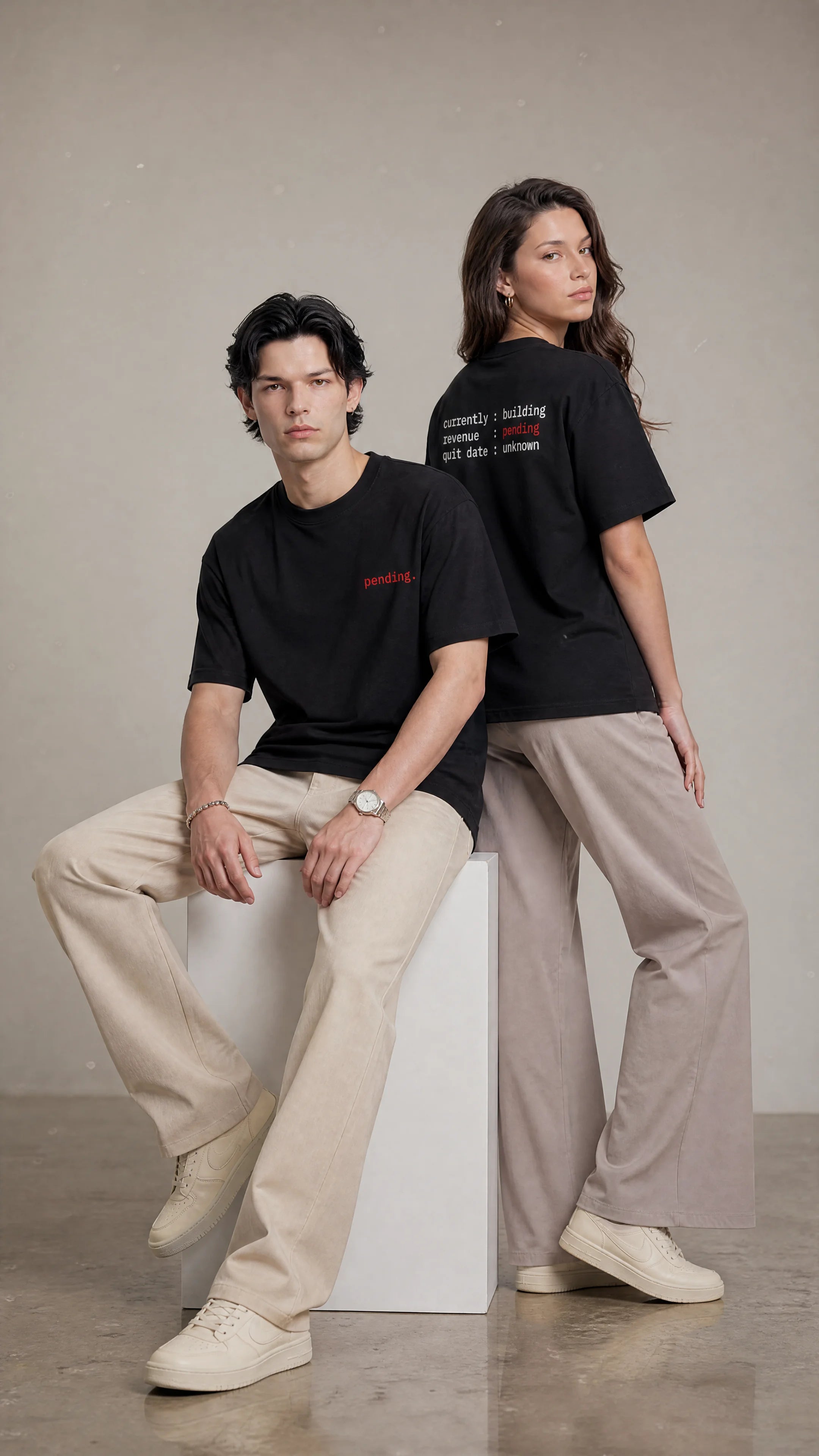 Two people wearing black t-shirts and beige pants sitting on a white block against a neutral background.