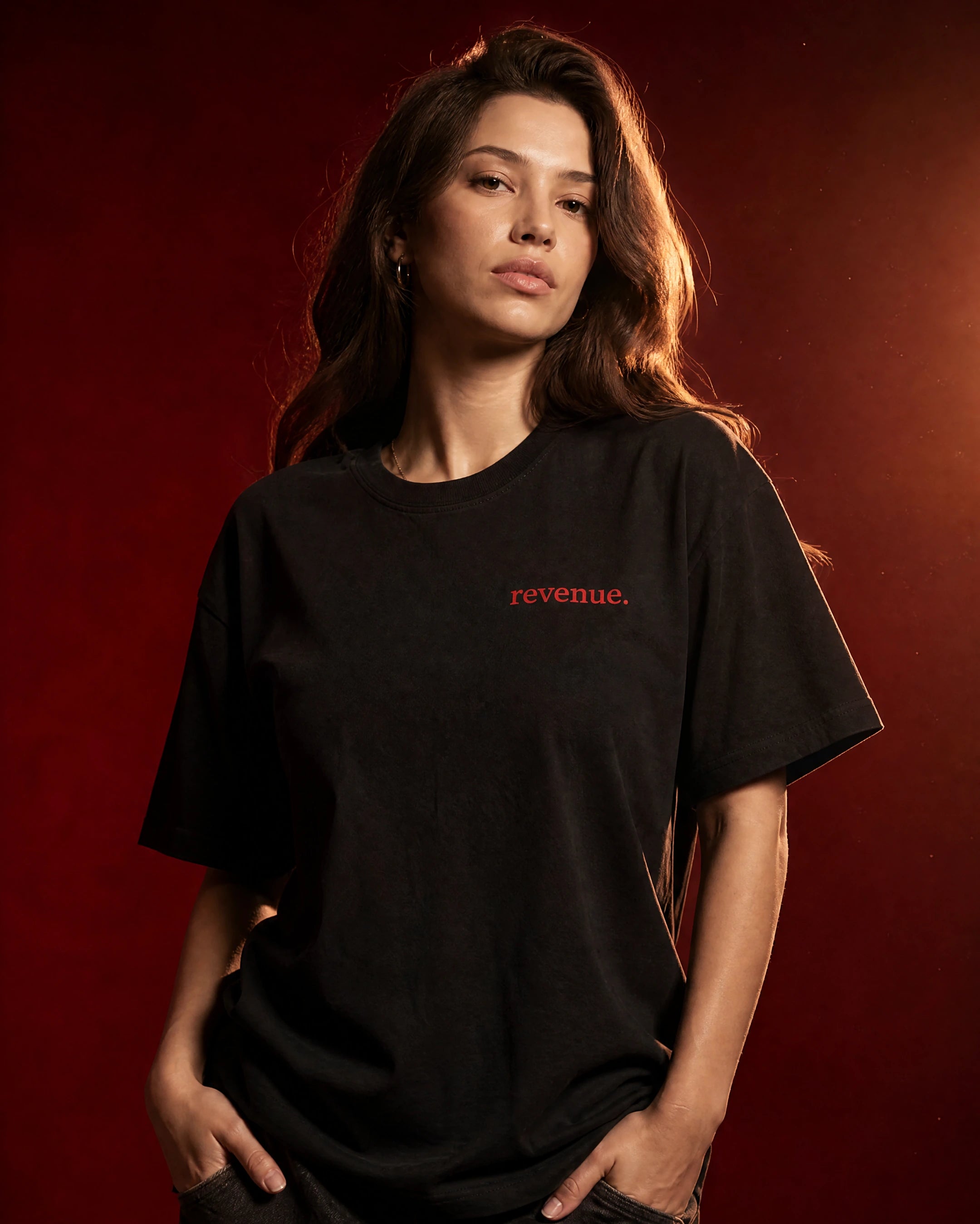 A woman modeling a black short-sleeve t-shirt with the word 'revenue.' printed in red on the left chest, set against a dark red studio background.