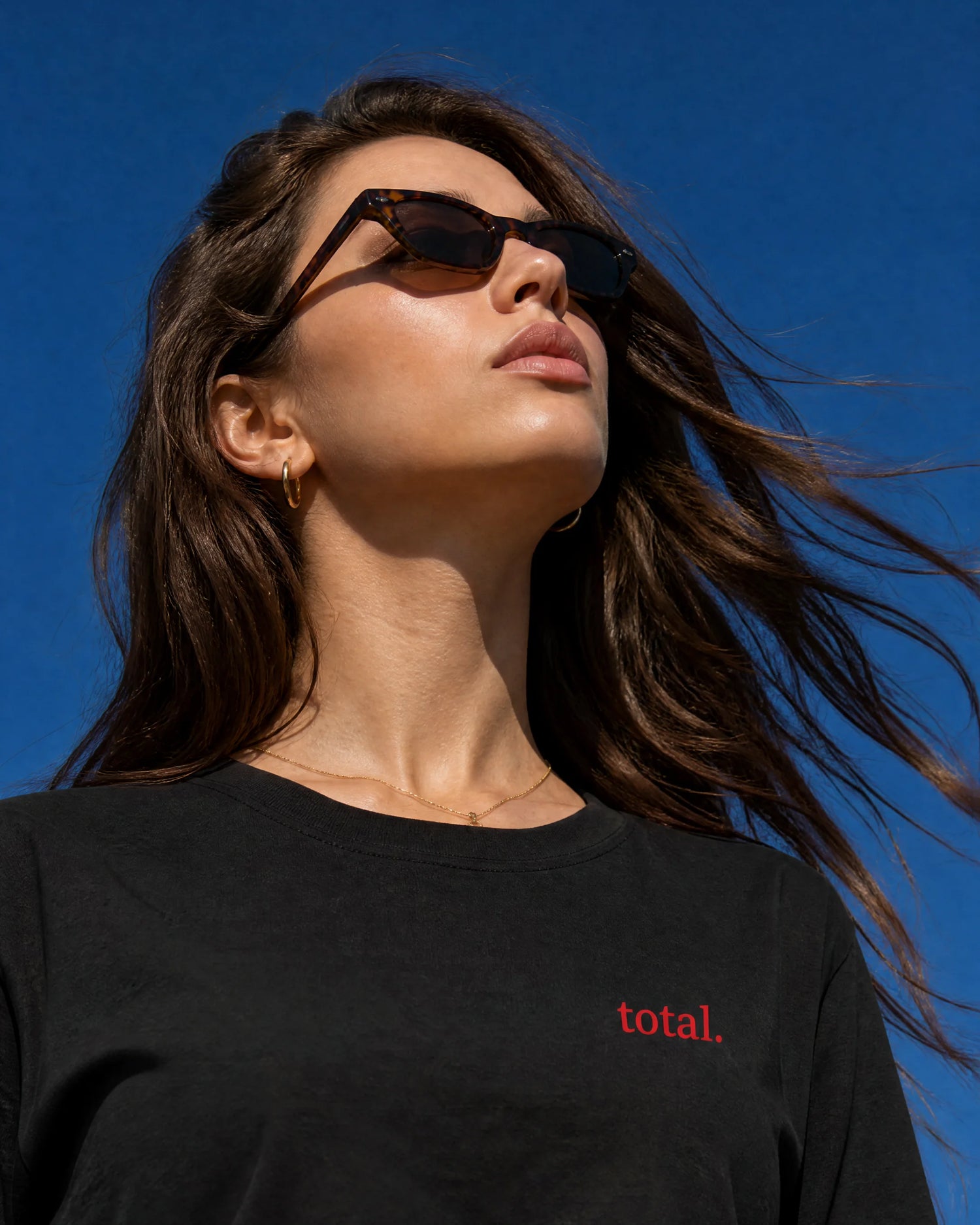 A woman wearing black sunglasses and a black t-shirt with the word 'total.' printed in red on the chest, photographed outdoors against a clear blue sky.