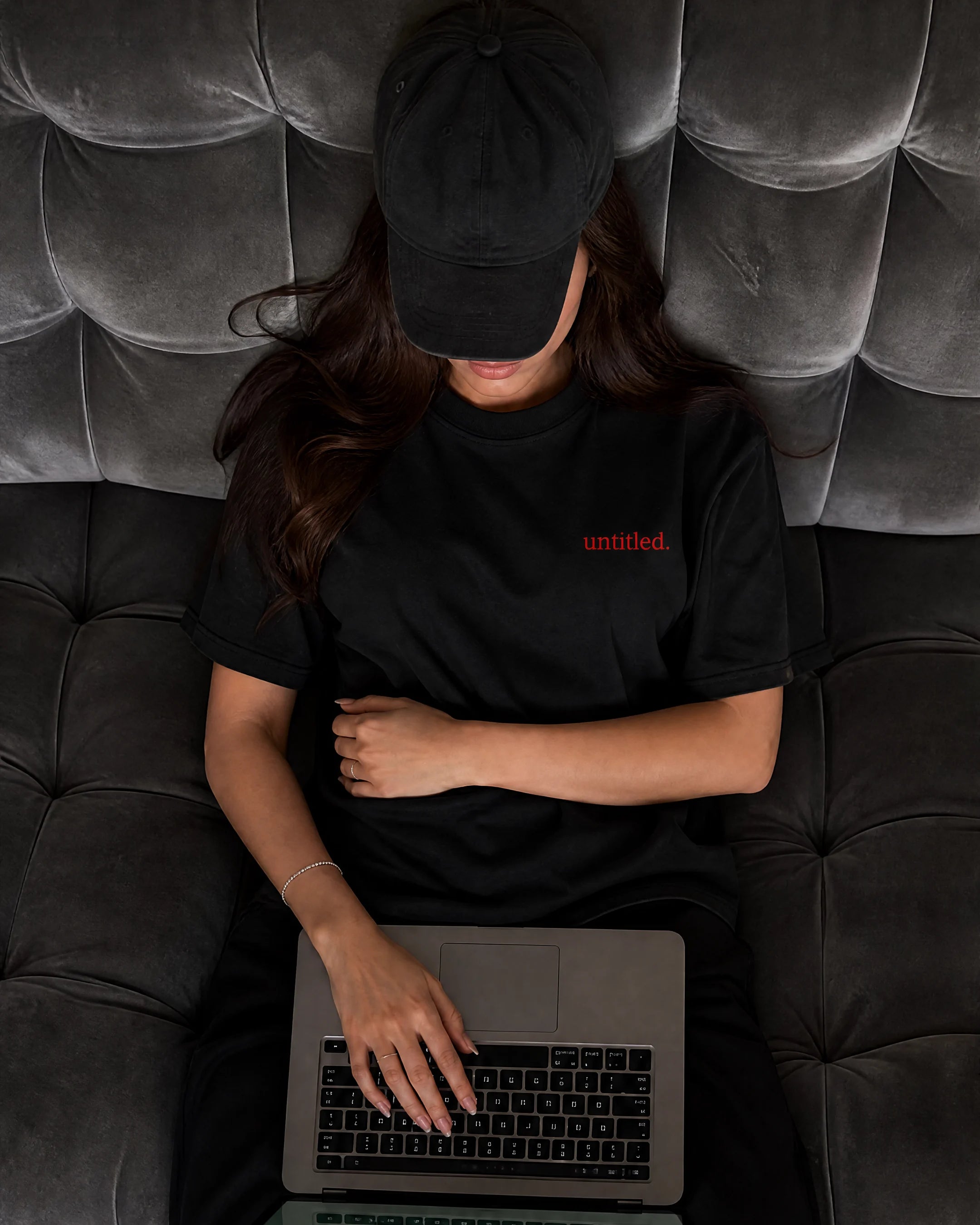Zehra wearing a black t-shirt with a small red 'untitled.' text on the chest, a black baseball cap, and black pants, sitting on a dark tufted leather couch and using a laptop.