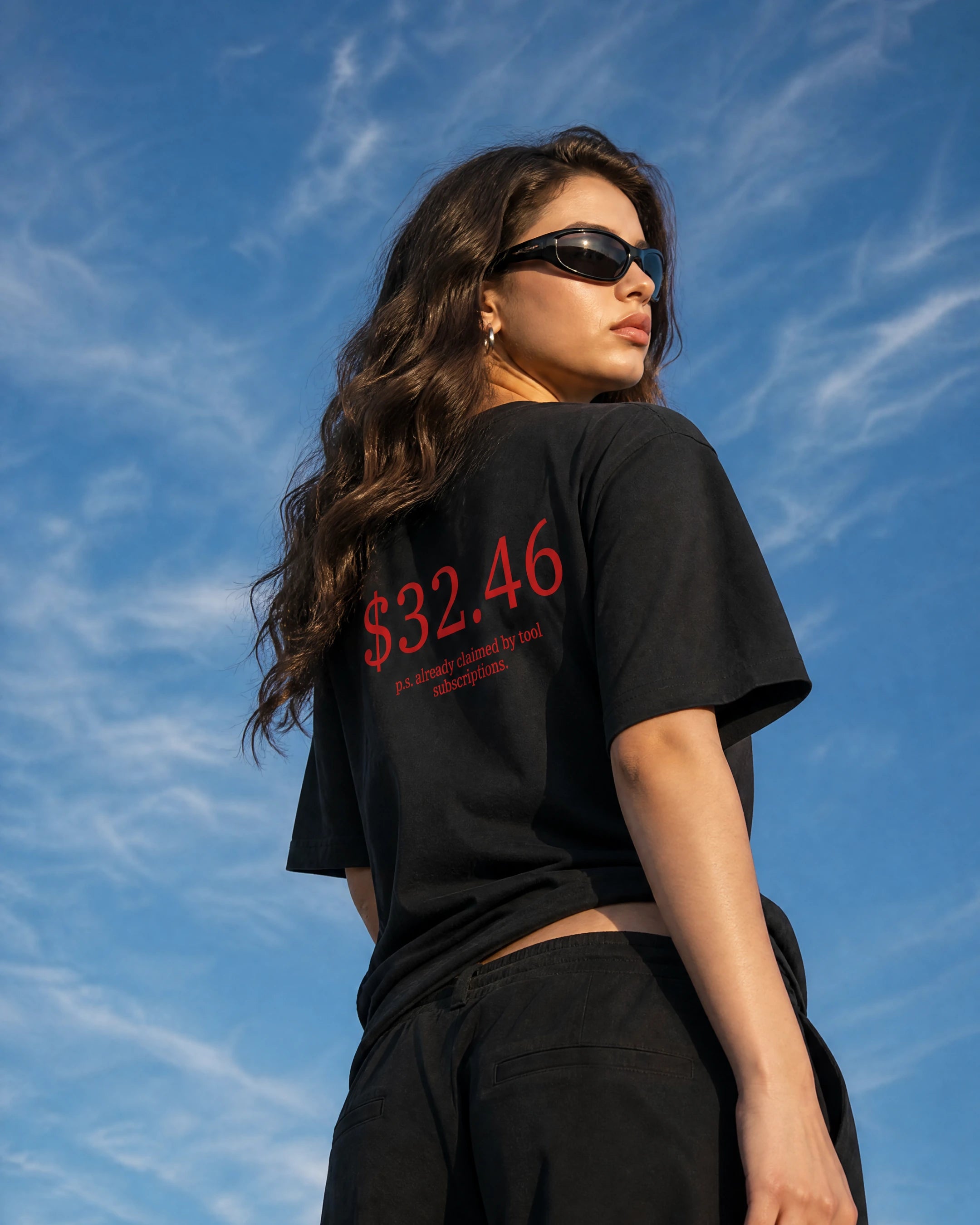 A woman wearing a black oversized t-shirt with red text reading '$32.46' and black sunglasses, photographed from a low angle against a blue sky with wispy clouds.