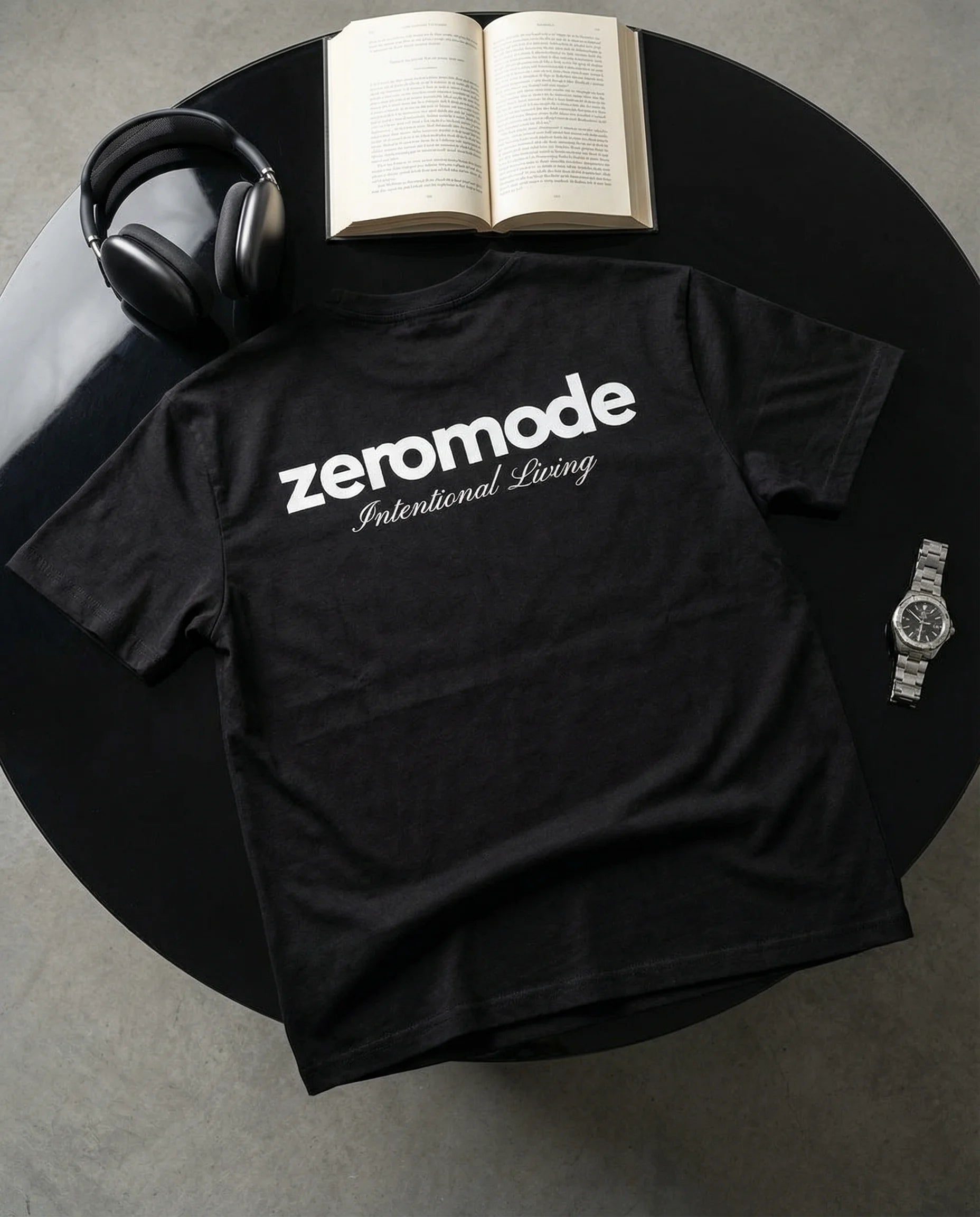 Black t-shirt with 'zeromode Intentional Living' text on a round table with a book and headphones.
